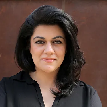 A woman with shoulder-length dark hair and a black top poses against a brown background. She has a neutral expression.