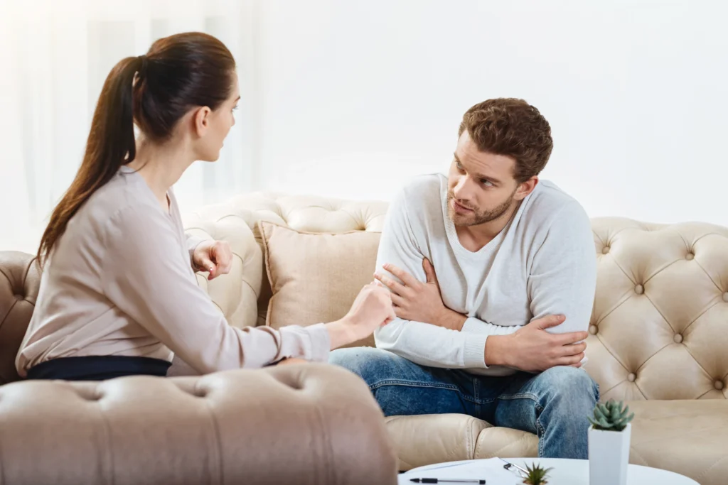 Two people, a woman and a man, sit on a beige sofa facing each other in conversation. The man has his arms crossed while listening to the woman, who gestures with her hand.