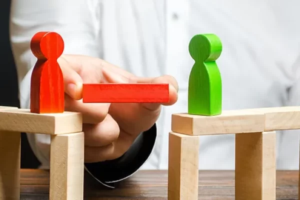 A person places a red block between a red and a green figurine, positioned on separate structures, symbolizing connection or bridging a gap.